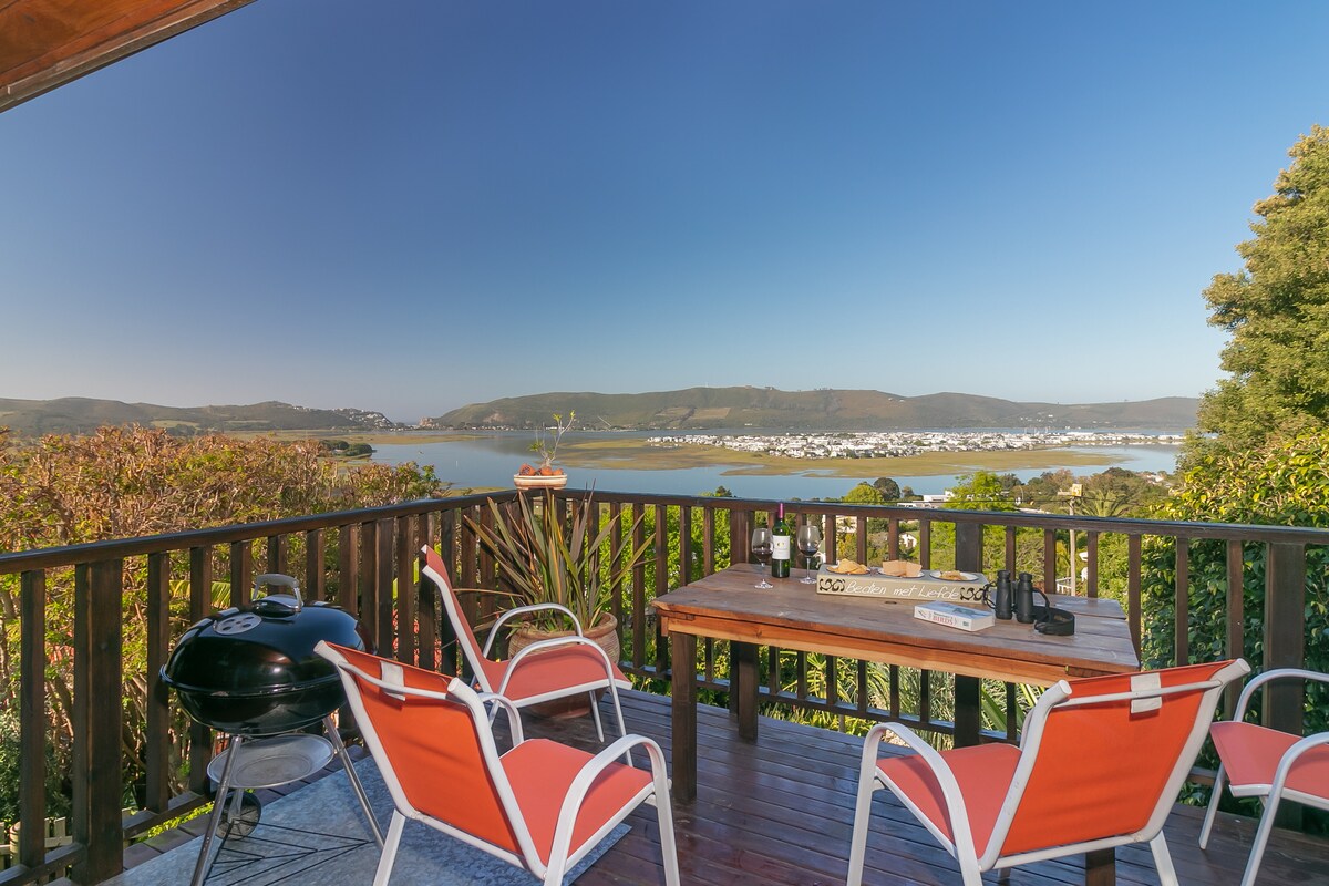 A spacious outdoor deck features a wooden table with seating for six, complemented by vibrant orange chairs. A barbecue grill stands nearby, offering scenic views of the surrounding landscape and water. Clear skies and distant hills enhance the serene atmosphere.