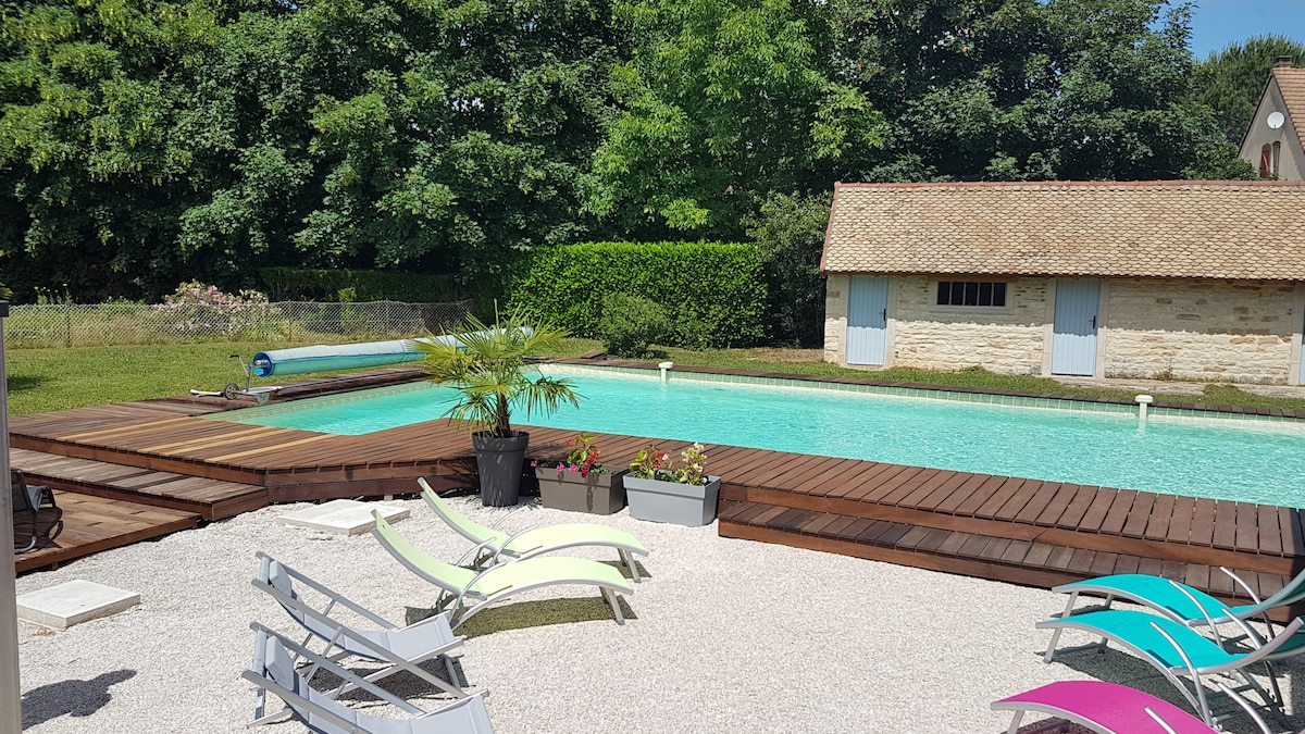 A tranquil outdoor space showcases a swimming pool surrounded by wooden decking. Sun loungers in various colors are arranged nearby, providing areas to relax. Lush greenery borders the pool, enhancing the serene atmosphere. A charming stone building is visible in the background.