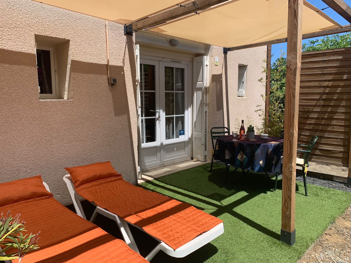 A private terrace features two loungers with orange cushions, surrounded by lush greenery. A small round table is set for two, offering a relaxed setting. Sunlight filters through a canopy overhead, enhancing the inviting space accessed through glass doors.