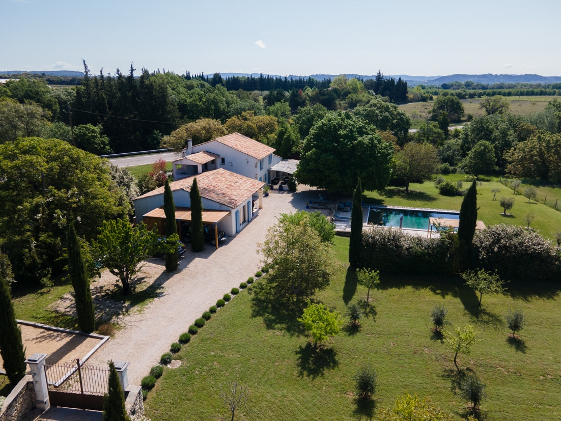 An aerial view captures a spacious property with a two-story house surrounded by lush greenery. A pristine pool is visible near the home, accompanied by well-maintained gardens and tall cypress trees dotting the landscape.