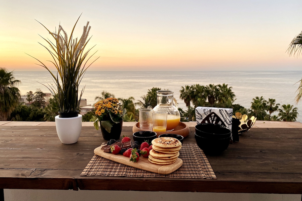 A wooden table is adorned with a spread of breakfast items, including pancakes, strawberries, and a glass pitcher. A tranquil ocean view and palm trees are visible in the background, illuminated by the warm hues of sunrise. Potted plants add a touch of greenery.