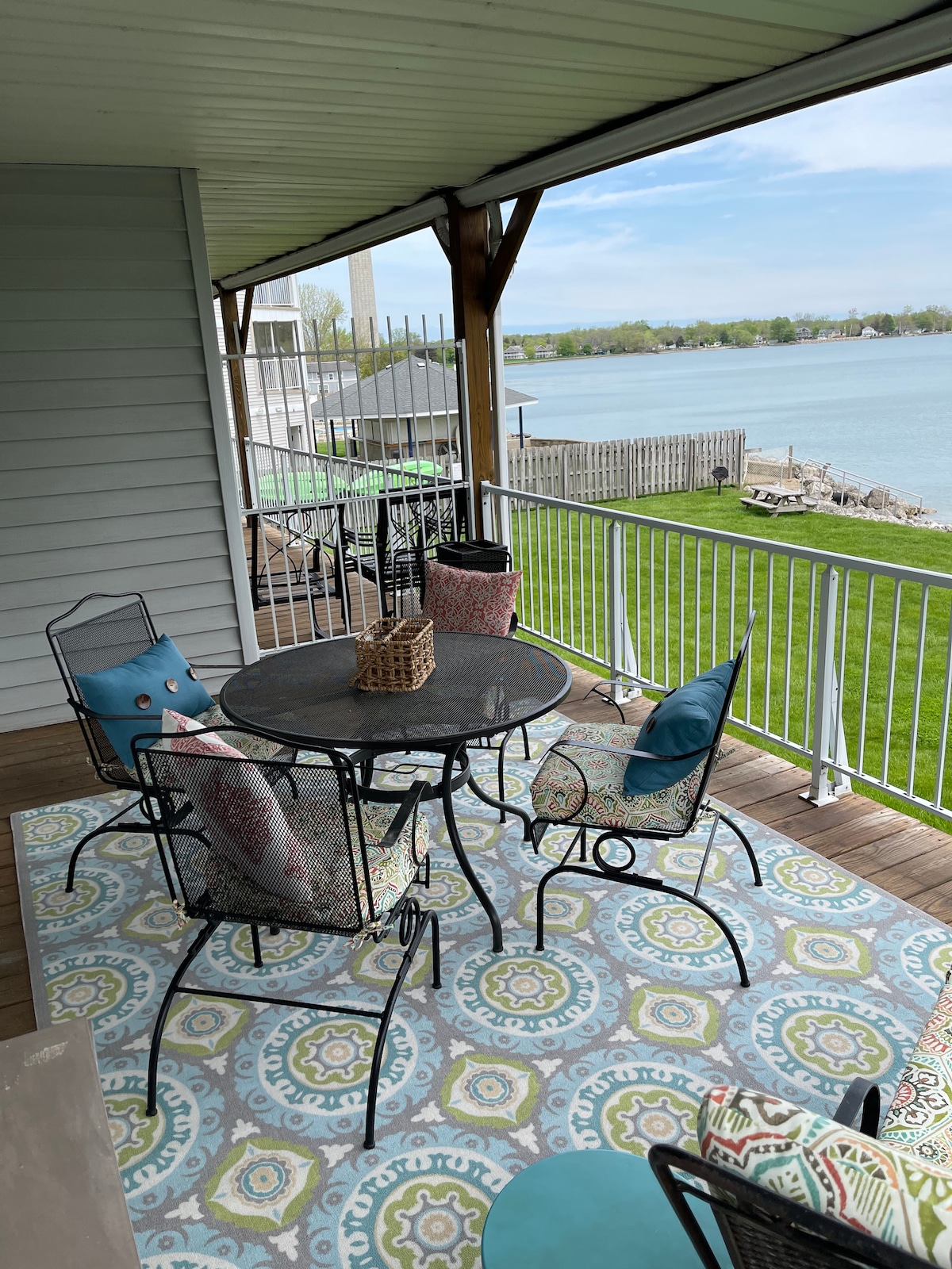 An outdoor deck features a round table surrounded by four iron chairs with cushions. A decorative rug adds color beneath the table, and a picturesque view of the lake is visible in the background alongside manicured grass and a wooden deck.