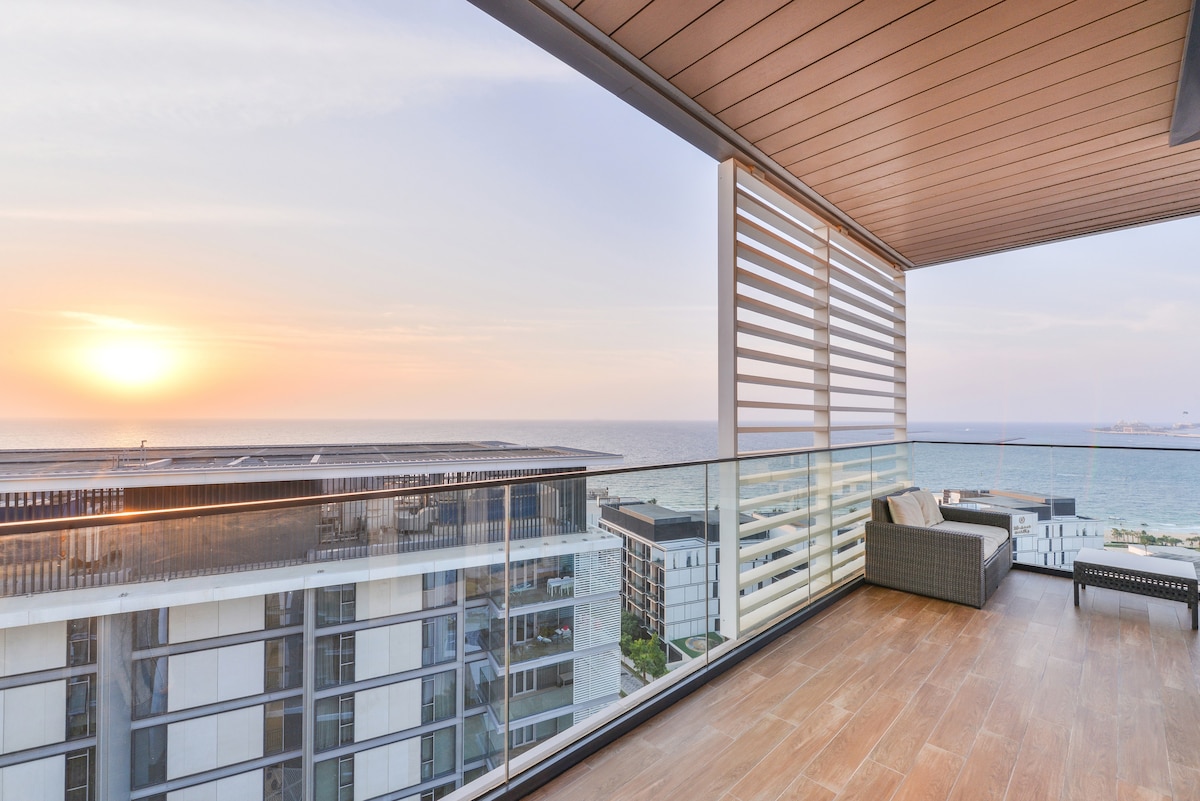 A spacious balcony offers stunning views of the Arabian Sea at sunset. The outdoor area features modern furnishings, including a cozy seating arrangement, framed by glass railings that capture the surrounding scenery.