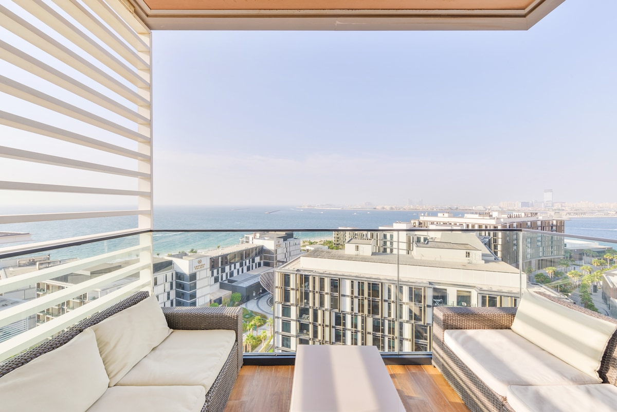 A furnished patio area features comfortable seating with plush cushions arranged around a sleek coffee table. The glass railing provides unobstructed views of the Arabian Sea, while soft sunlight illuminates the space, creating a serene spot for relaxation.