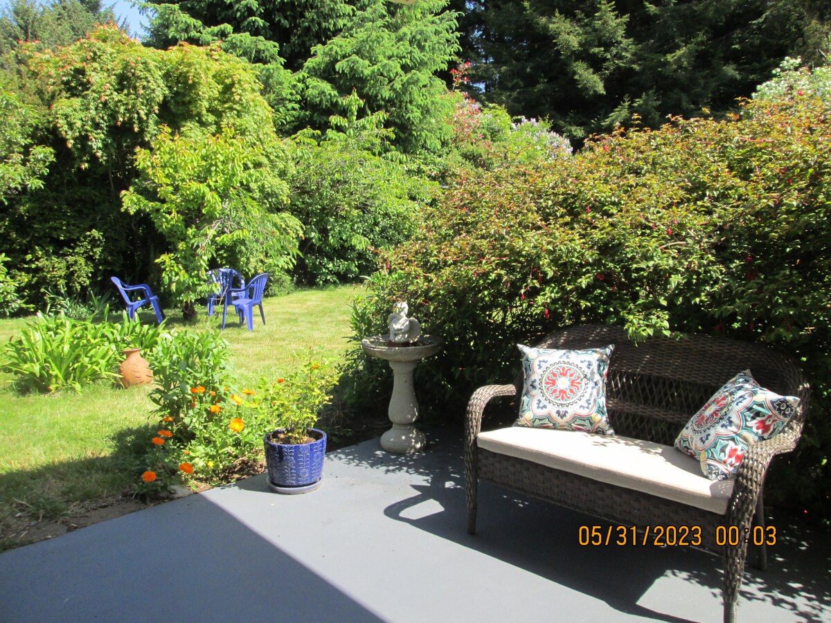 A shaded outdoor area features a comfortable wicker bench with patterned cushions, positioned near a small flower pot. A stone fountain stands nearby, while vibrant greenery surrounds the space, and two blue chairs are visible in the grassy background.