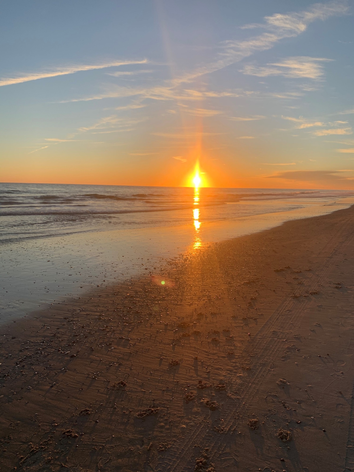 A sunset view casts warm golden hues over the ocean, reflecting on the wet sandy beach. Gentle waves roll in, and a serene atmosphere is created by the vast open sky, which contains soft clouds trailing into the horizon.