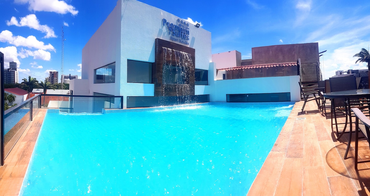 A large swimming pool is presented, featuring clear blue water and a waterfall feature. Surrounding the pool are lounge chairs and an open area with a view of the sky, enhancing the leisure space. The building's facade is visible in the background.