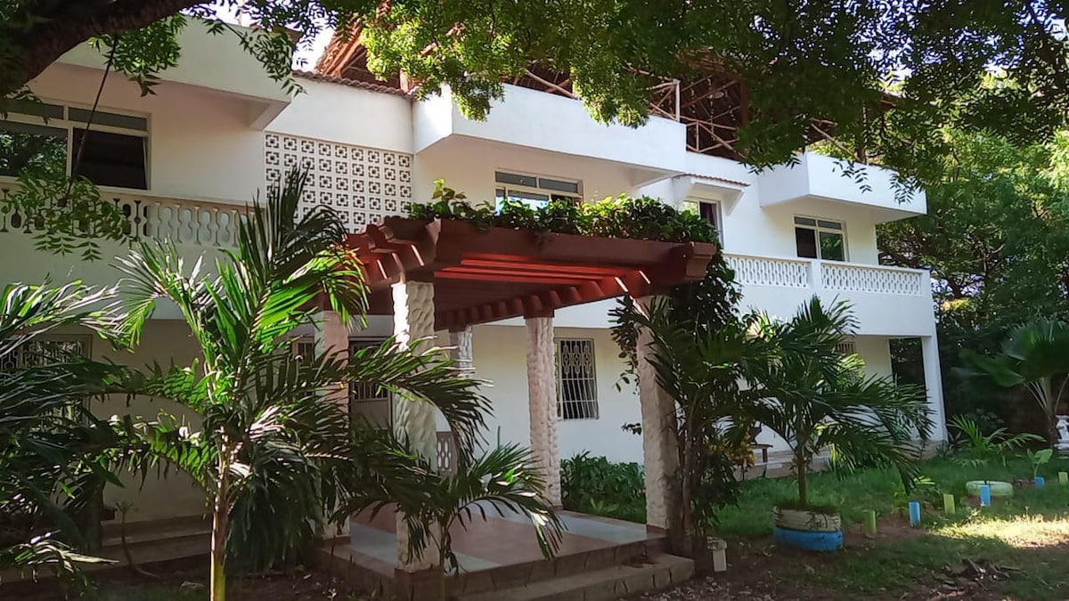 The exterior showcases a multi-level building framed by lush greenery. A welcoming entryway features a wooden pergola adorned with climbing plants. The surrounding area includes well-maintained gardens and palm trees, providing a serene and inviting atmosphere.