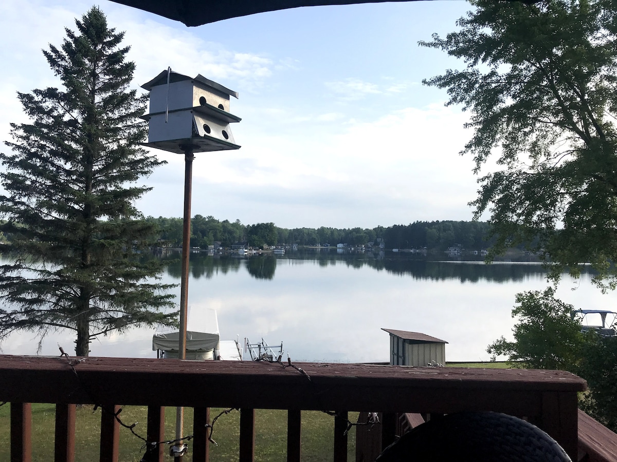 A serene view of the lake is framed by a wooden deck railing, featuring a birdhouse and a tall pine tree in the foreground. The calm water reflects the surrounding greenery and distant homes, creating a peaceful atmosphere.