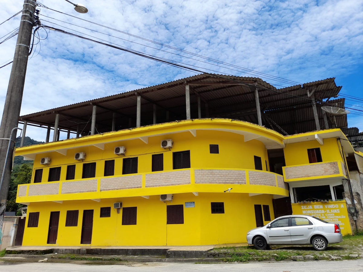 A two-story yellow building with multiple air conditioning units is depicted. The structure features a covered terrace and a wooden roof under construction. A parked white car is visible in front, with a backdrop of blue sky and scattered clouds.