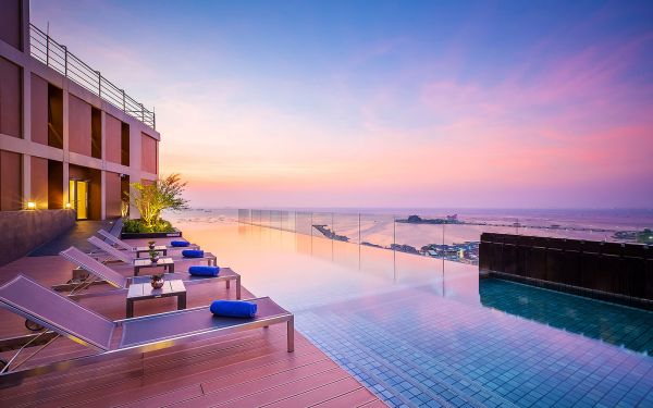 An outdoor infinity pool is nestled on a rooftop, with lounge chairs positioned nearby. The tranquil water reflects the soft colors of the sunset sky, merging pastel hues of pink and purple. City and coastal views are visible in the background, promoting a relaxing atmosphere.