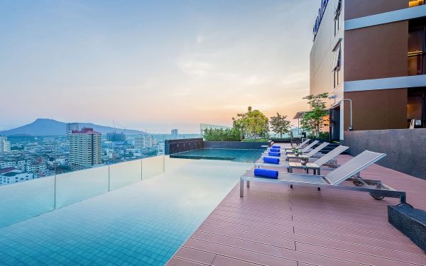An outdoor infinity pool is featured on a rooftop terrace, offering expansive views of the surrounding cityscape and mountains. Lounge chairs with blue cushions are positioned near the pool's edge, set against a tranquil evening sky illuminated by soft pastel hues.