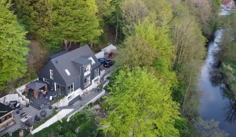 An aerial view captures a modern house nestled among lush greenery, adjacent to a winding river. The property features multiple outdoor areas with seating, while vehicles are parked nearby. The surrounding trees create a serene natural backdrop.