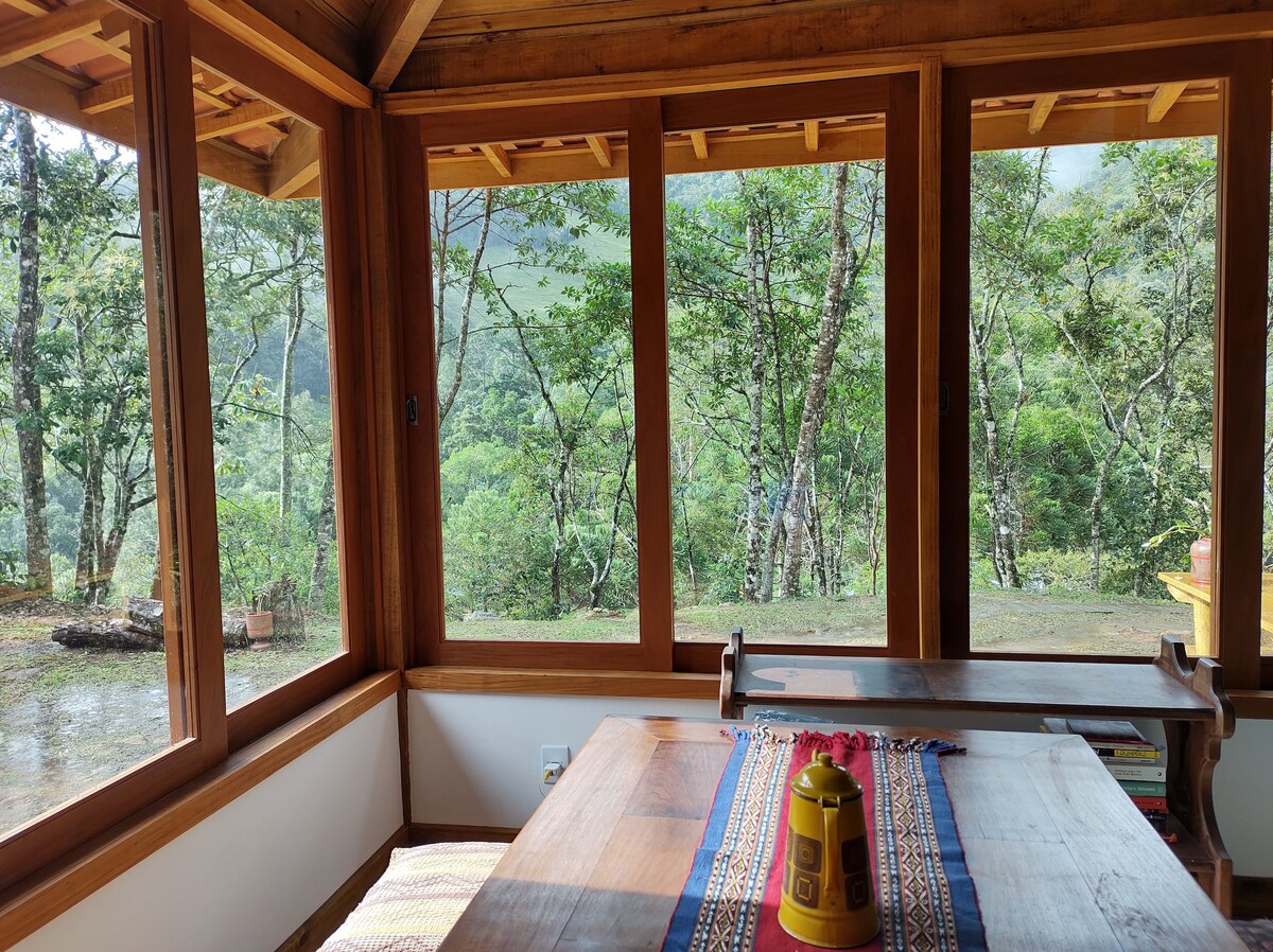 A bright dining area features large windows that provide an expansive view of the surrounding trees and hillside. A wooden table is set with a decorative runner and a yellow container. Natural light fills the space, creating a connection with the outdoor landscape.