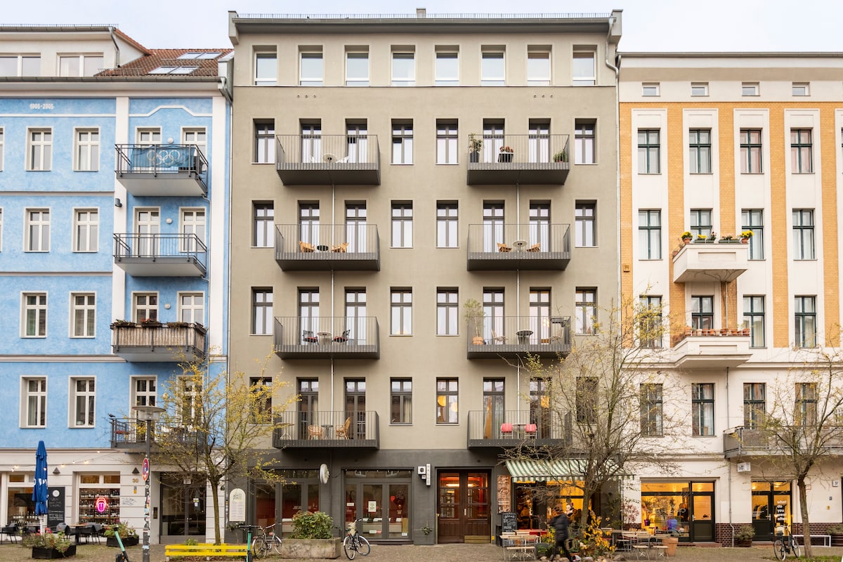 The exterior of a classic Berlin residential building from 1898 is showcased, featuring a neutral facade with balconies. Various window styles and a cafe on the ground floor contribute to the lively street atmosphere. Well-maintained trees line the sidewalk, enhancing the urban environment.