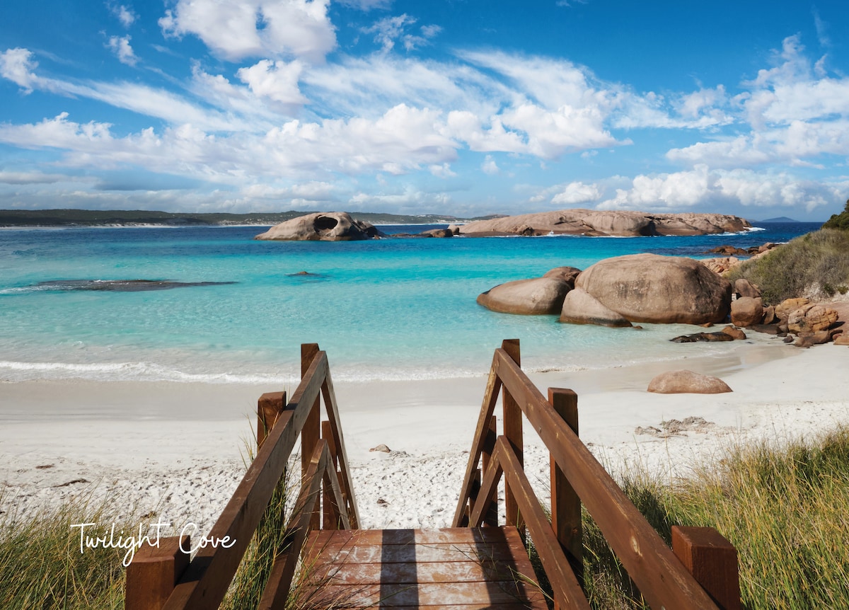 A wooden walkway leads down to a serene beach, with soft white sand and clear turquoise waters. Large rock formations are visible in the distance under a partly cloudy blue sky, creating a tranquil coastal setting.