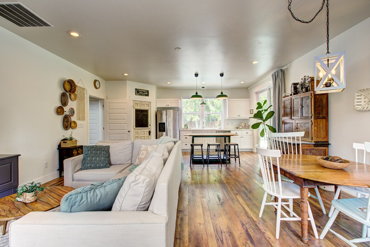 The open-concept living area showcases a spacious lounge with a light-colored sectional sofa, accompanied by a dining table set for six. Modern pendant lights illuminate the space, which features a blend of natural wood flooring and white cabinetry.