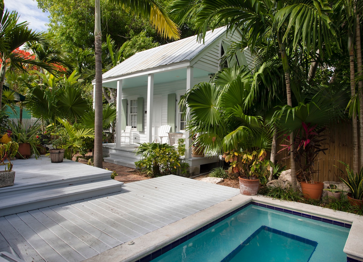 A charming white house is surrounded by lush tropical landscaping, featuring various palm trees and vibrant foliage. The elevated porch is framed by white rocking chairs. A tranquil pool sits beside a wooden deck, providing a serene outdoor space.