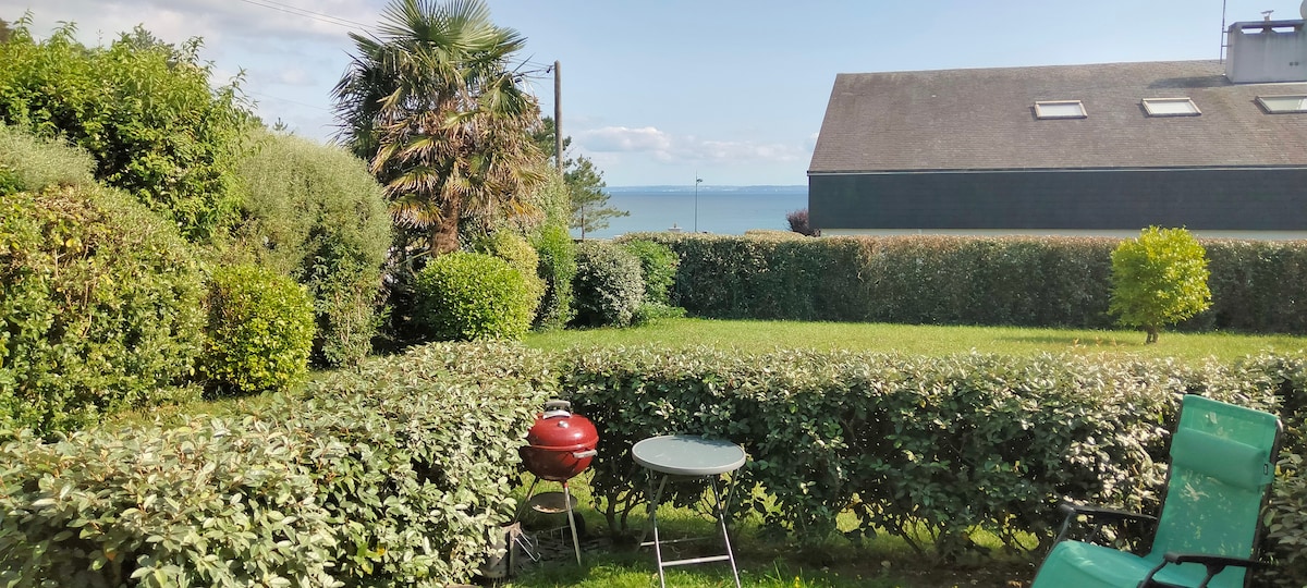 A well-maintained garden area features neatly trimmed hedges and is equipped with a grill and a small table with chairs. The view beyond includes the sea in the distance and a clear blue sky above, providing a serene outdoor space.