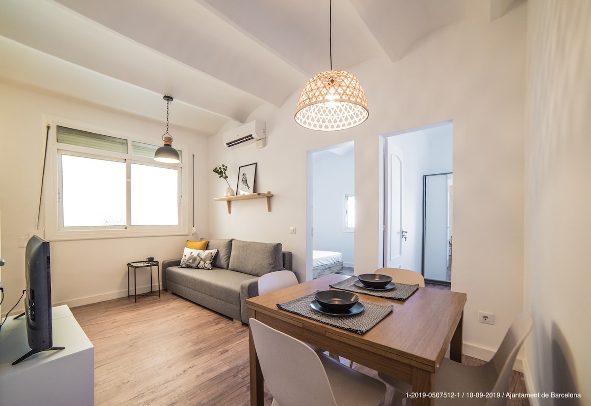 A bright living area features a comfortable grey sofa, a wooden dining table set for two, and a wall-mounted television. Natural light filters through the window, creating a warm ambiance. Minimalist shelving adds functional decor, while the overall design emphasizes a fresh, modern aesthetic.