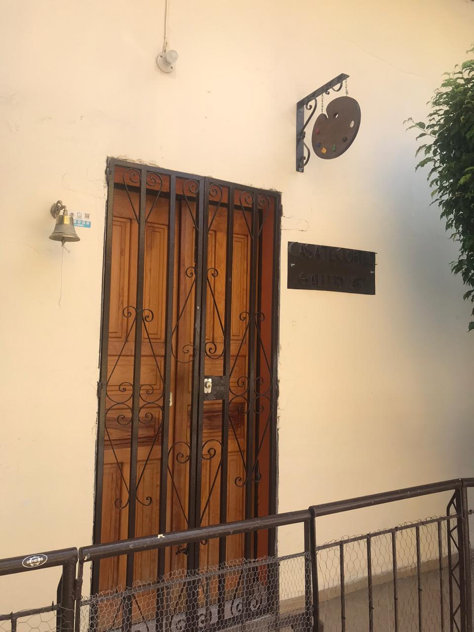 A wooden door with decorative iron bars is framed by a light-colored wall. A small bell is located next to the door, while a sign hangs above, indicating 'Galería 67.' A potted plant is positioned nearby, adding a touch of greenery to the entrance.