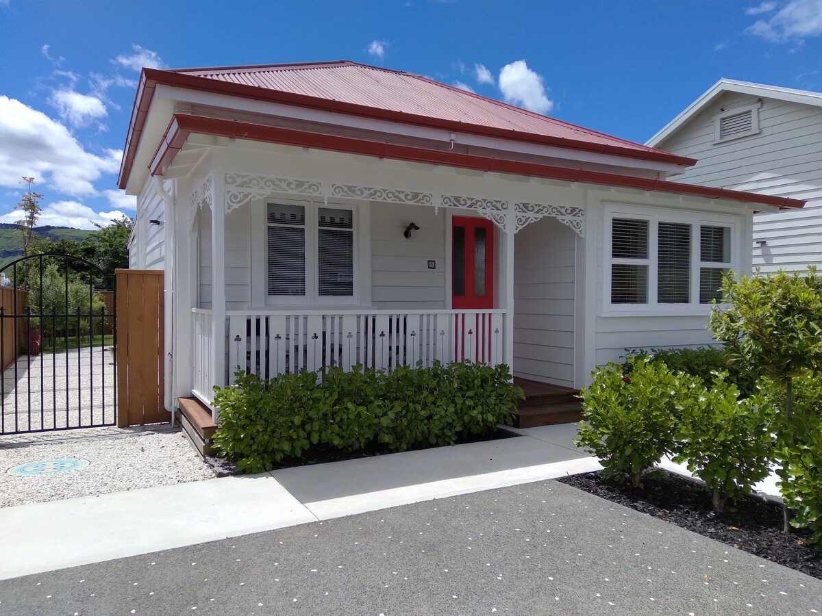 The charming exterior of a railway cottage is displayed, featuring a red-trimmed roof and a bright red front door. A welcoming porch with decorative trim leads to a manicured garden and gravel driveway, surrounded by low greenery and a gated entrance.