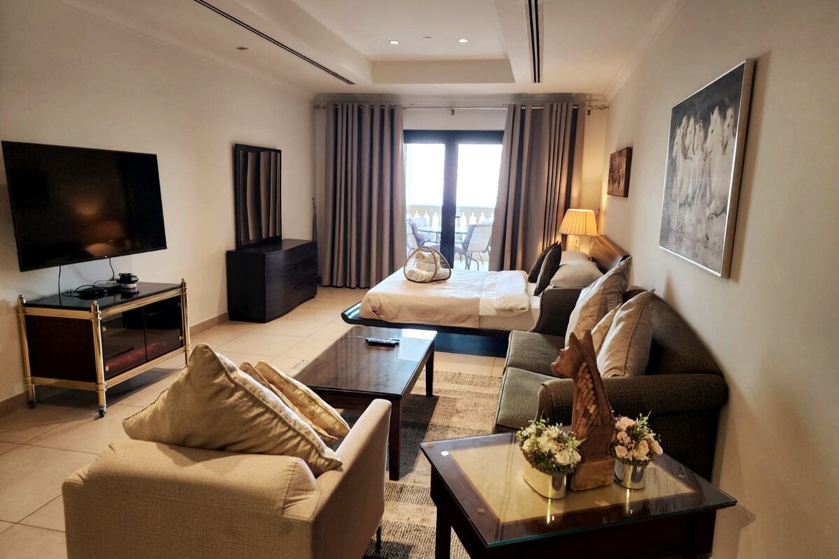 The spacious living area is furnished with a comfortable sofa set and an elegant coffee table. A flat-screen TV is mounted on the wall, and ample natural light fills the room through large windows leading to the balcony, creating a bright and inviting space.