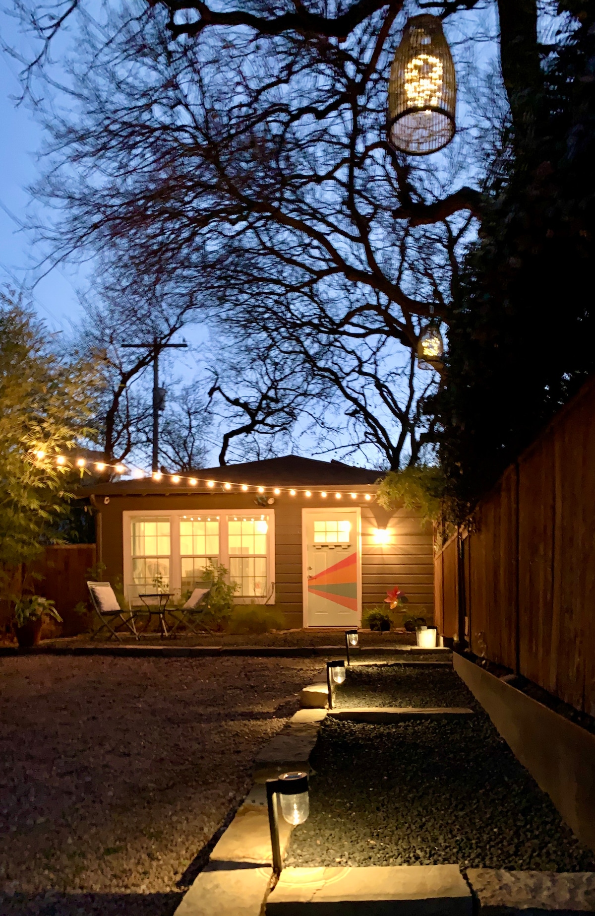 An inviting exterior view of the studio is captured during twilight, showcasing warm light emanating from the windows. String lights are draped above a gravel path leading to the private entry, framed by trees and greenery for a serene ambiance.