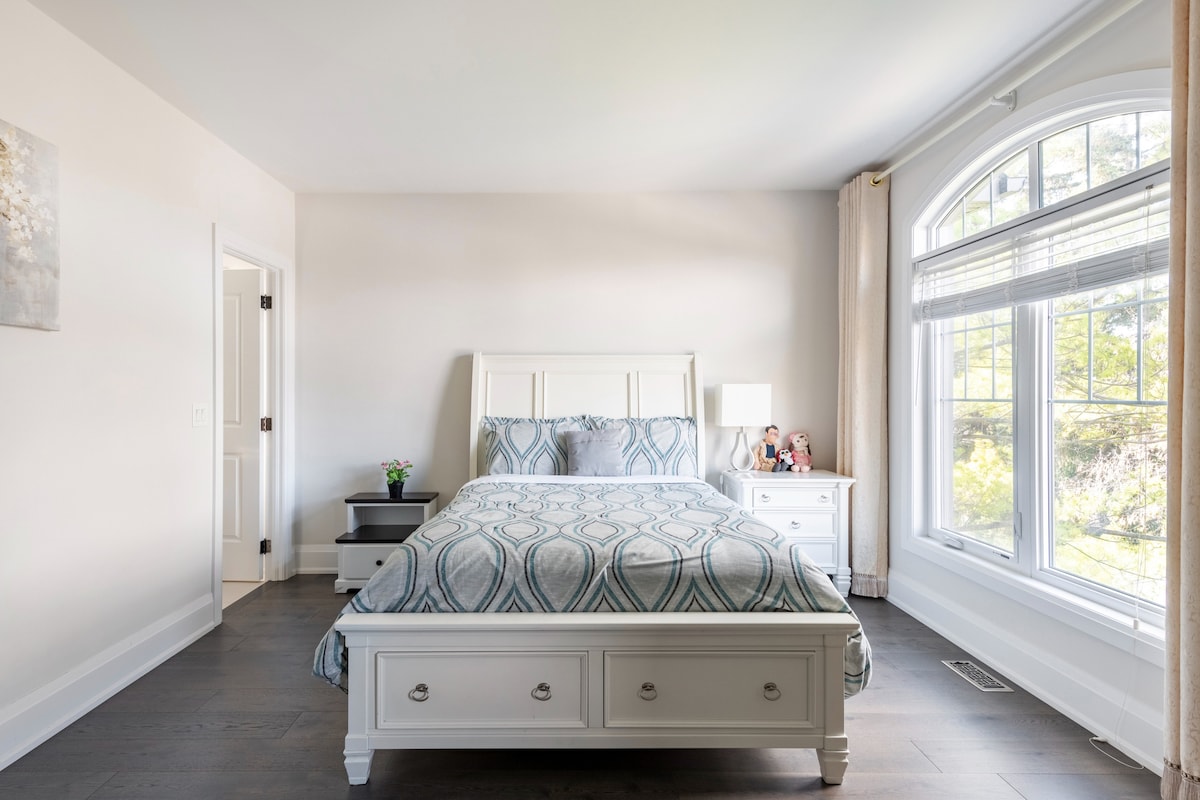 A bright bedroom features a queen-size bed with a patterned comforter. Soft natural light enters through a large, arched window, illuminating the pale walls. A bedside table with a lamp is positioned next to the bed, while a small plant adds a touch of greenery to the room.