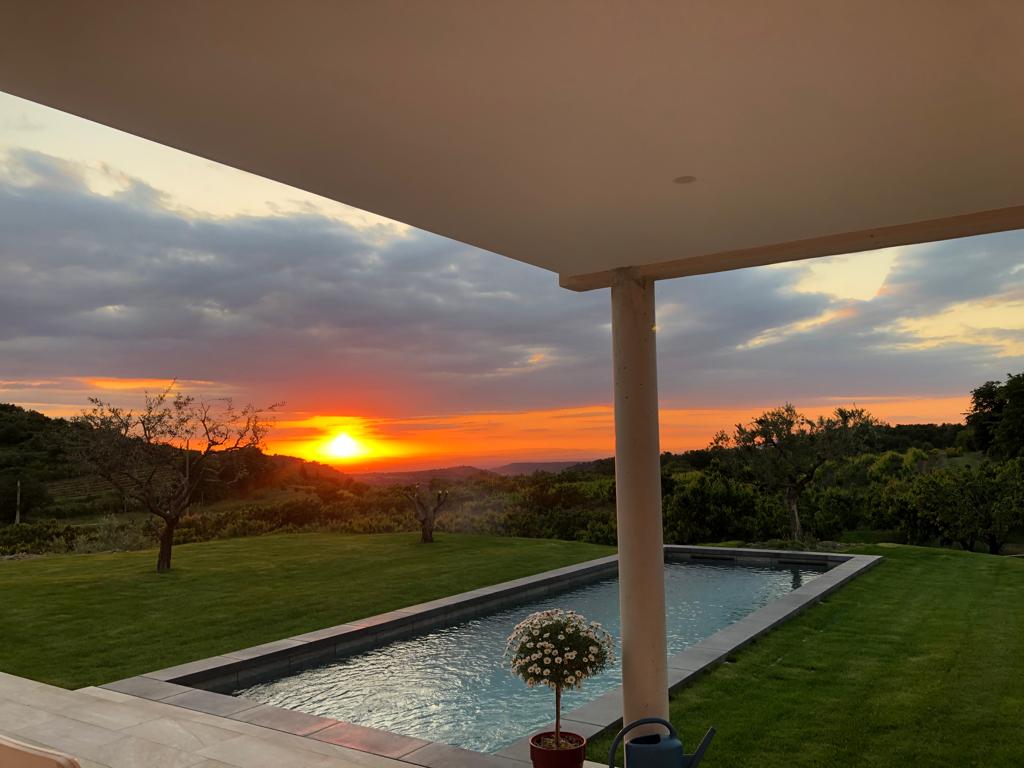 A serene outdoor setting showcases a heated swimming pool beside a spacious green lawn. The sun sets in the background, casting warm hues across the sky. A flower pot is positioned near the poolside, enhancing the natural beauty of the landscape.