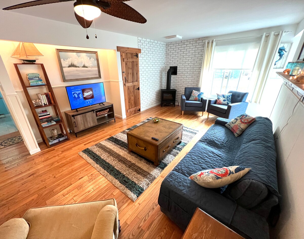 A cozy living room features light wood flooring and a striped area rug. A comfortable sofa and two armchairs provide seating, with a coffee table at the center. A wooden cabinet houses a TV and books, while a decorative piece rests on the shelf, creating a welcoming atmosphere.