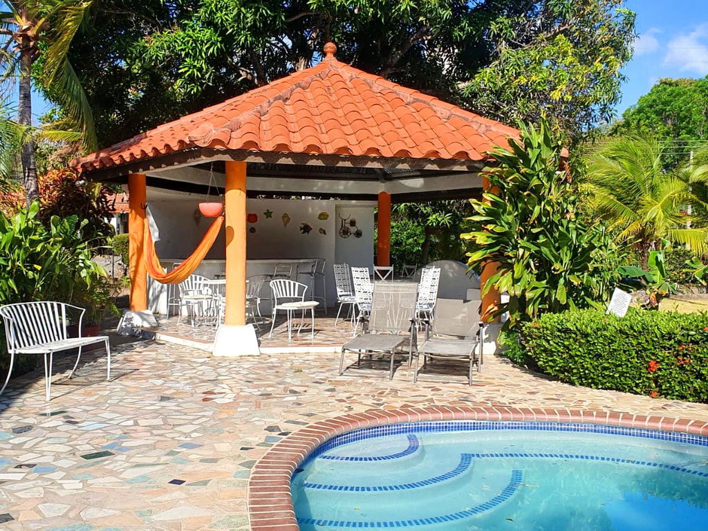 A shaded outdoor gazebo features a tiled roof, surrounded by tropical greenery. Equipped with multiple lounge chairs and a dining area, it offers a comfortable space for relaxation. A small pool is situated nearby, enhancing the serene atmosphere of the outdoor area.