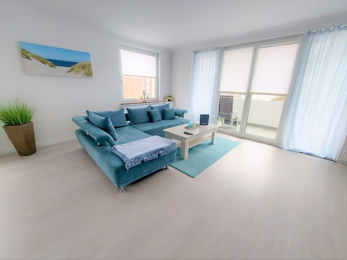 A spacious living room features a light blue sofa accompanied by a matching area rug and a coffee table. Large windows allow natural light to fill the space, with soft white curtains filtering the view. A coastal artwork enhances the ambiance, complemented by a potted plant.