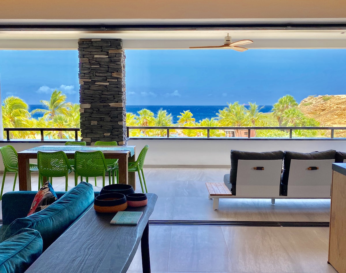 A spacious living area features modern furnishings, including a comfortable sofa and a dining table with vibrant green chairs. Large windows offer unobstructed views of the Caribbean Sea and surrounding palm trees, creating a bright and airy atmosphere.