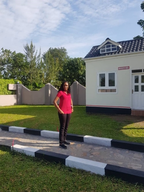 A pathway bordered by contrasting black and white markings leads to a white building with a pitched roof. Lush green grass surrounds the area, and trees are visible in the background, creating a natural setting. The structure features a welcoming entry door and a minimized window.