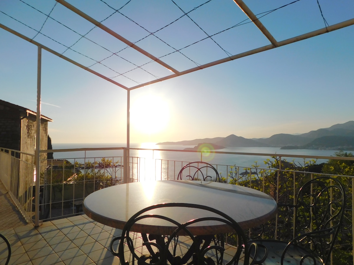 A balcony is presented with a round table and four chairs, offering an unobstructed view of the sun setting over the sea. Light reflects off the water, creating a serene backdrop against the mountainous coastline.