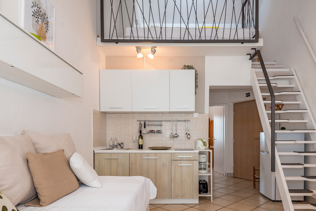 A bright and functional kitchen area is visible, featuring modern cabinetry and essential appliances. A cozy seating space is present, with light-colored cushions laid on a white sofa. A spiral staircase leads to an upper gallery area, allowing access to additional sleeping or reading space.