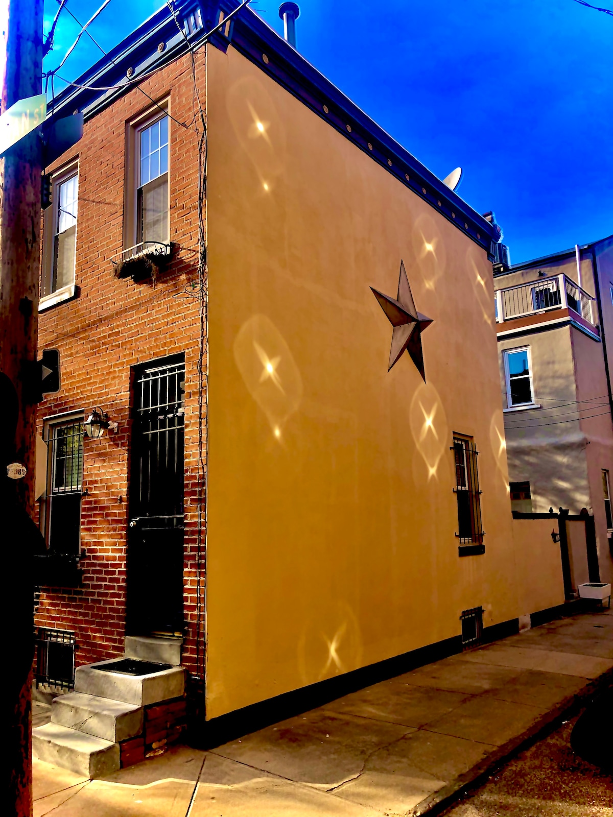 The exterior of the trinity home is presented, showcasing a yellow wall adorned with a decorative star. Natural light creates playful reflections on the surface. The image captures the building’s unique structure on a tree-lined street, emphasizing the charm of the neighborhood.