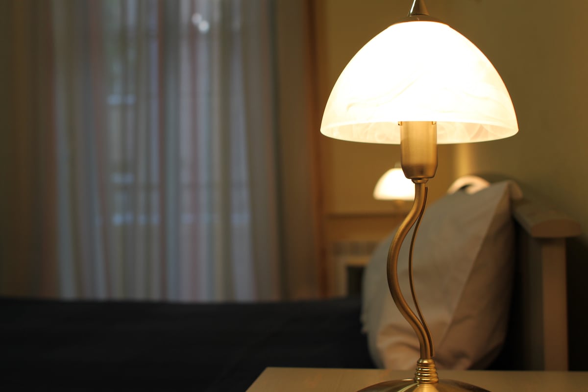 A bedside lamp with an elegant design is illuminated, casting a soft glow over the nightstand. The background features a neatly made bed with a white headboard and soft linens, while sheer curtains filter light from a nearby window.