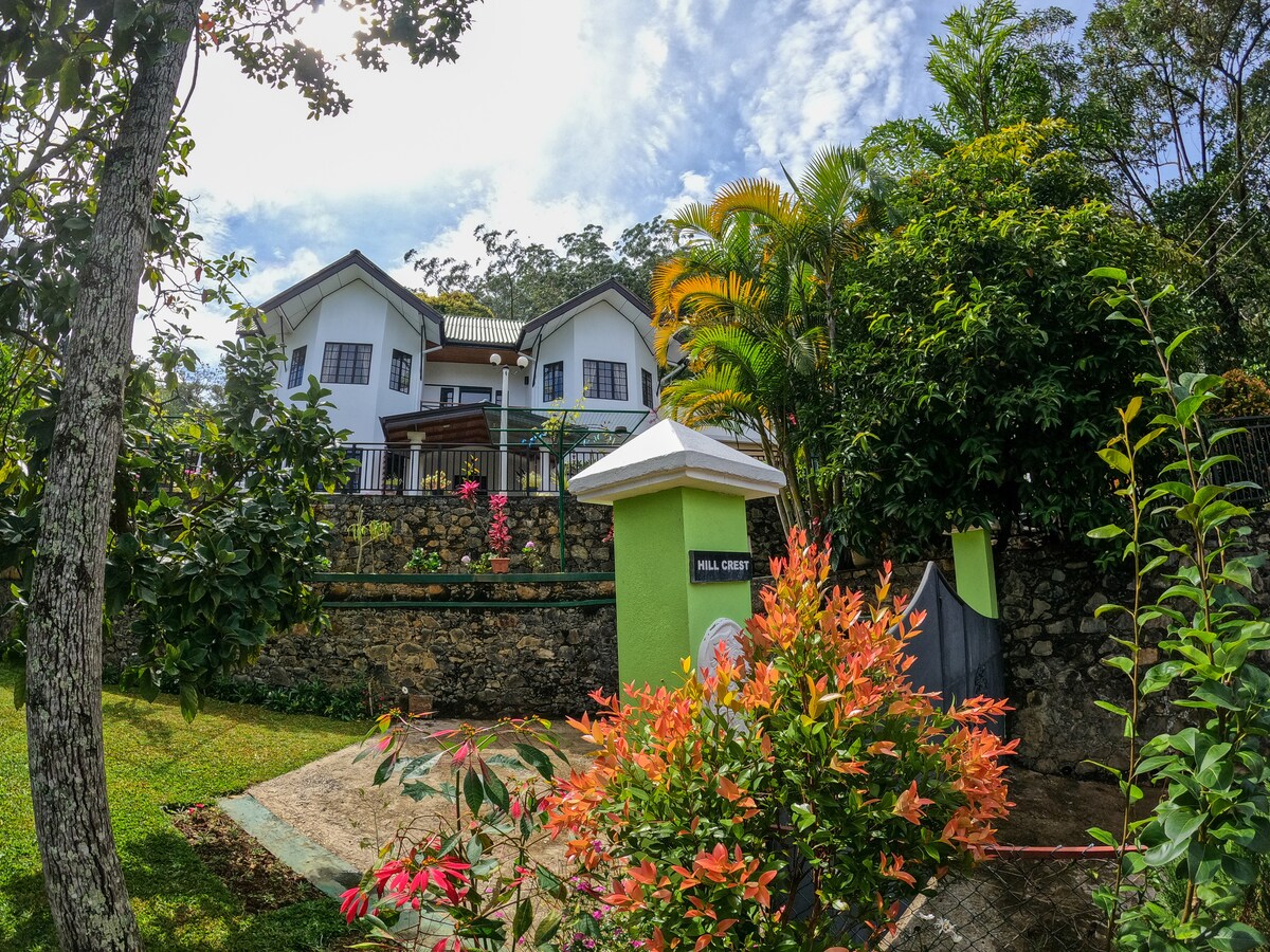 The exterior of the bungalow is surrounded by lush greenery and colorful flower beds. A garden area is visible in the foreground, while a two-story structure rests against the backdrop of trees. A welcoming entrance with a decorative pillar is highlighted.