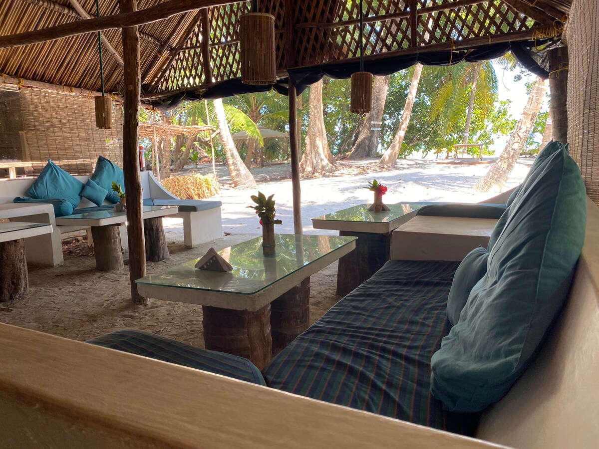A shaded seating area features comfortable blue cushions on white benches, inviting relaxation. Glass-topped tables are positioned among a sandy floor, alongside vibrant tropical plants. In the background, palm trees and distant beach scenery create a serene setting.