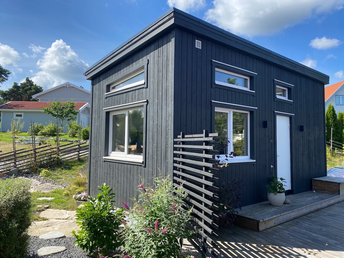 A modern, dark wooden guesthouse is situated amid greenery, featuring large windows that allow natural light to fill the interior. A small porch is visible at the entrance, surrounded by flower beds and a decorative wooden lattice, enhancing the inviting ambiance of the exterior.