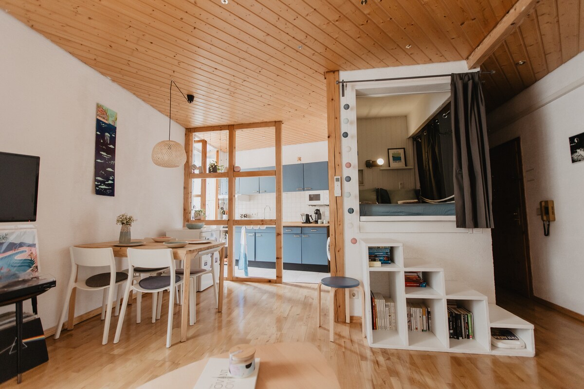 An open living area showcases a wooden ceiling and light-colored flooring. A dining table with four chairs is situated near a kitchen with blue cabinetry. A small bookshelf and decorative items add interest, while soft natural light filters through a nearby window.