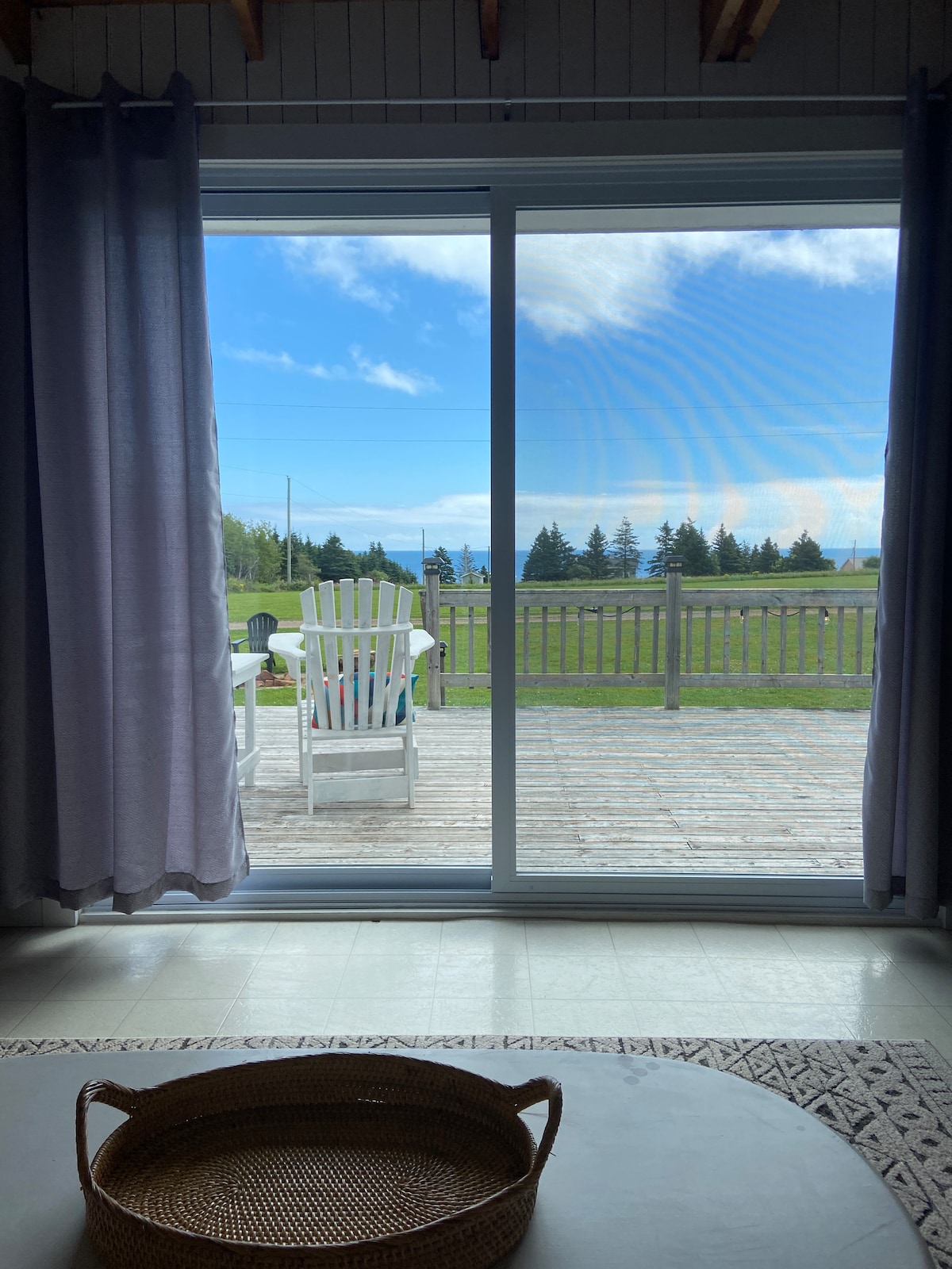 A sliding glass door offers a view of a large outdoor deck with a colorful adirondack chair, extending to a grassy area. The landscape features vibrant green grass and distant trees under a clear blue sky.