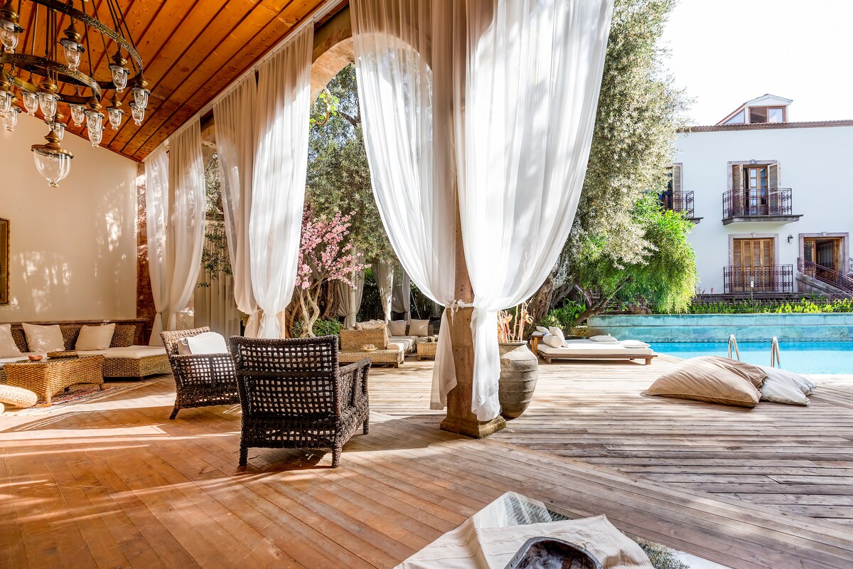 A spacious patio area features inviting seating arrangements with comfortable woven chairs and plush cushions. Sheer curtains gently sway in the breeze, allowing natural light to illuminate the wood decking. A serene swimming pool is visible in the background, surrounded by lush greenery.