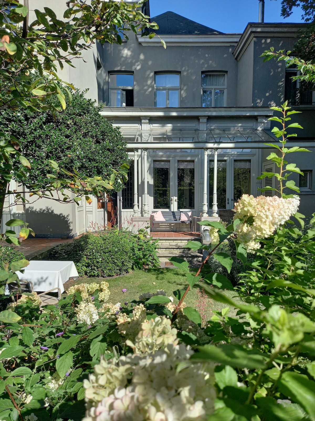 The serene garden setting features lush greenery and blooming flowers, with a glimpse of a classic home structure in the background. A spacious terrace with outdoor seating is visible, allowing for relaxation amongst the natural surroundings.
