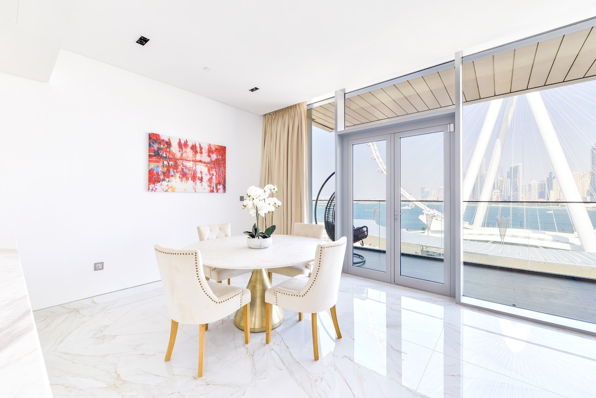 A bright dining area features a round table surrounded by four upholstered chairs. A colorful artwork adorns the wall, while large glass doors lead to a furnished terrace, providing views of the Arabian Sea and Ain Dubai.