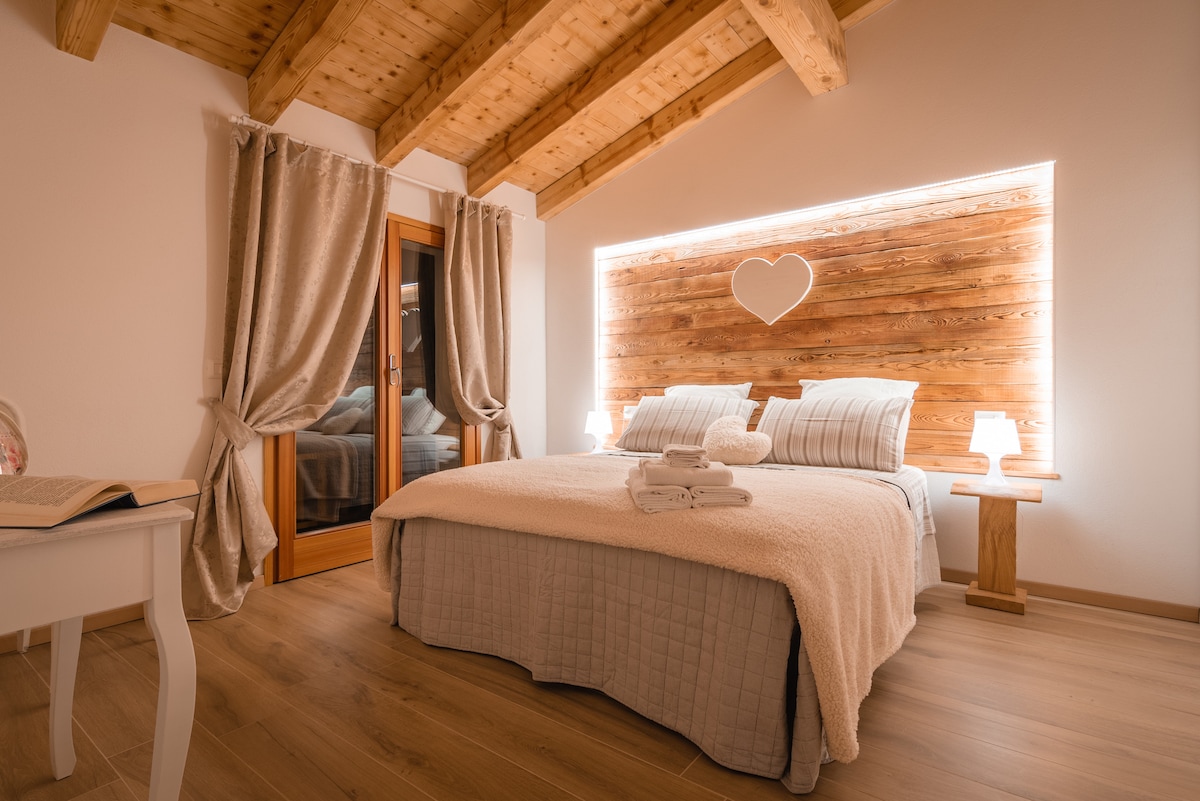 A cozy bedroom is highlighted by warm wooden accents on the walls and ceiling. A spacious bed is covered with a light blanket, while bedside tables with lamps provide soft illumination. Large windows are draped with curtains, allowing for natural light.
