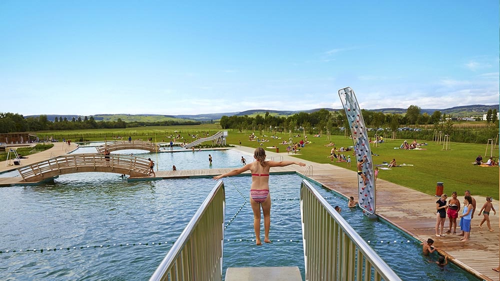A scenic outdoor area is displayed, featuring a person balancing on a platform above water. Lush green landscapes surround multiple swimming spots, with bridges and visitors relaxing on the grassy banks. A climbing wall is visible on the side, enhancing the recreational atmosphere.