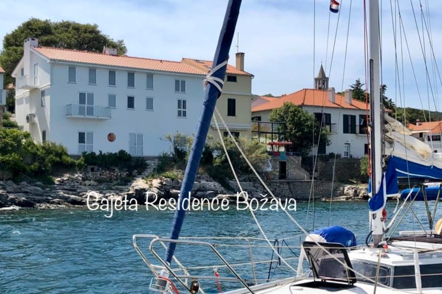 The Gajeta Residence Bozava is depicted from the water, showcasing its bright white facade with a terracotta roof. Nearby buildings are visible, framed by lush greenery. A sailboat is in the foreground, anchored in the clear waters, enhancing the coastal atmosphere.